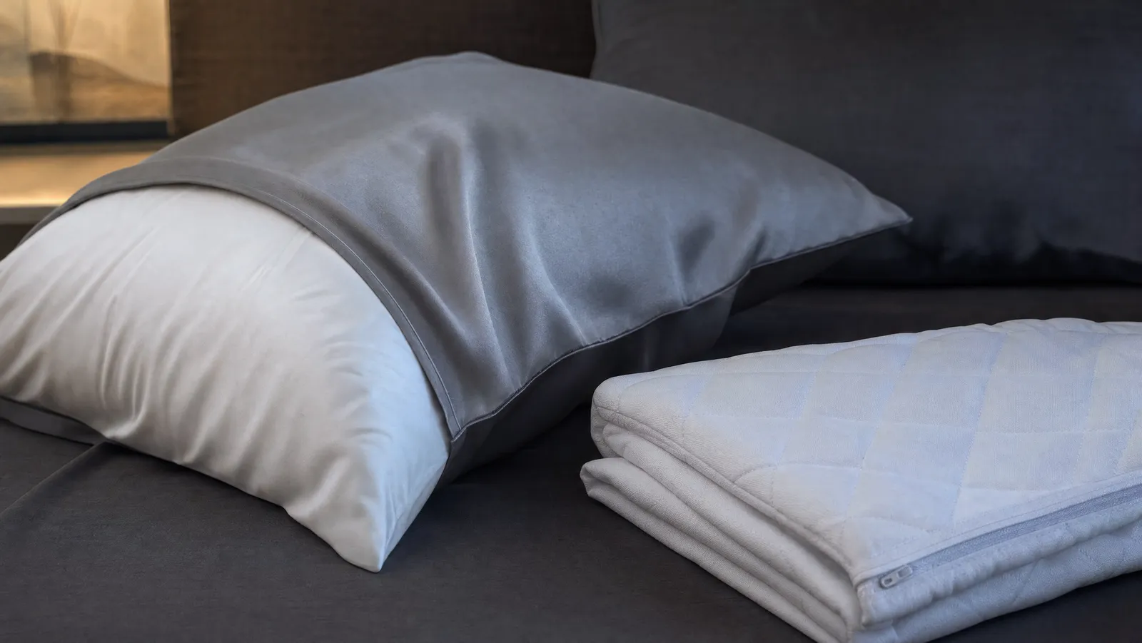 A thin silver-grey cooling pillowcase half pulled onto a white pillow on a dim bed, with a quilted waterproof pillow protector folded beside it