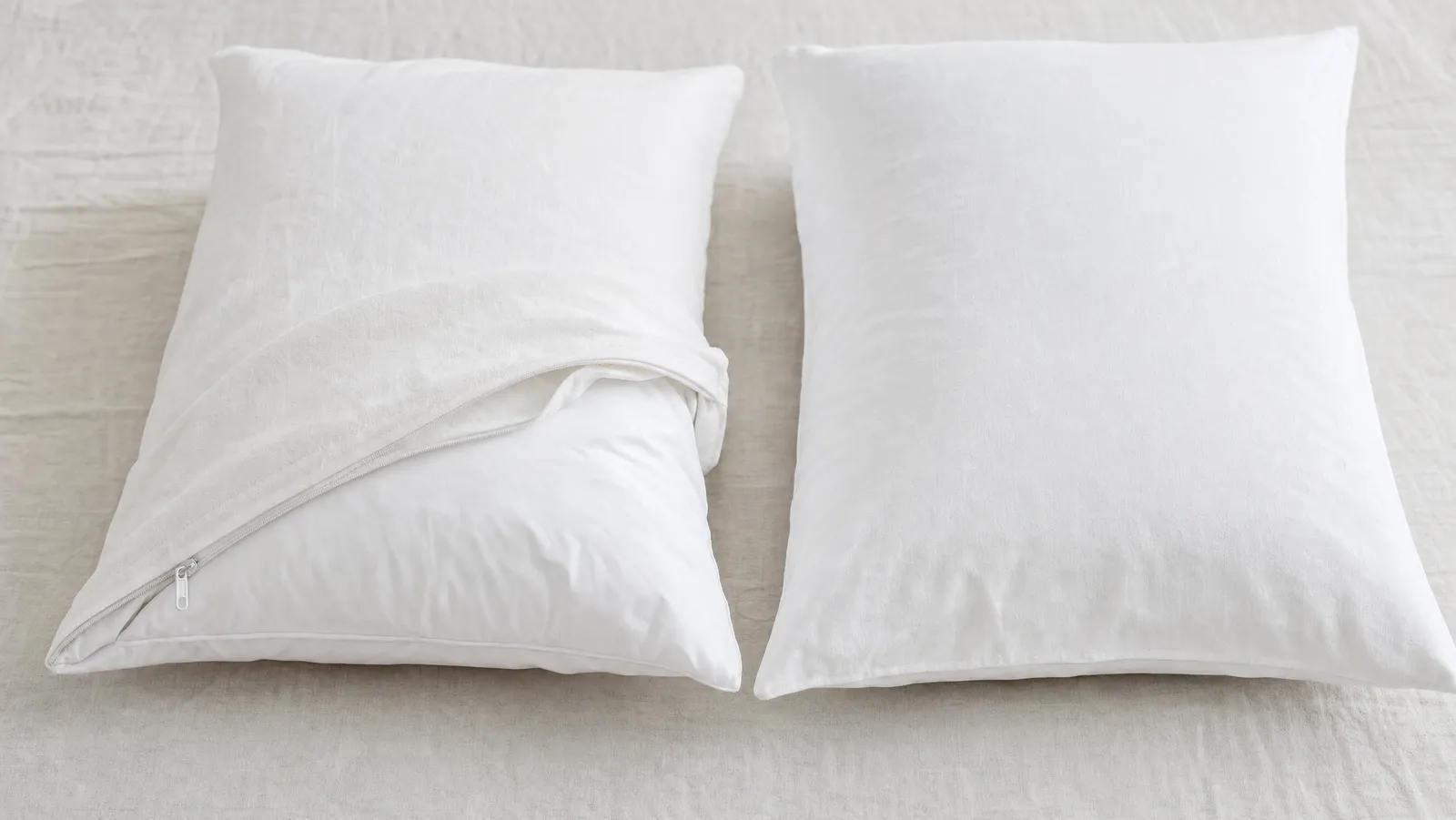 Two white bed pillows side by side on pale linen, one in a partly unzipped cotton protector revealing the pillow inside